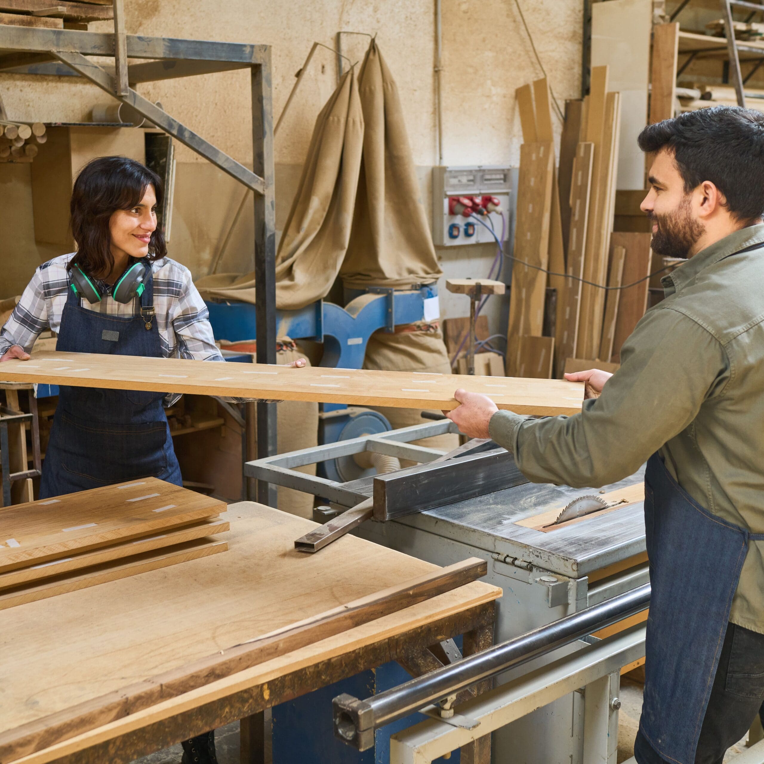 Two people working in a woodshop, holding a board between them and smiling.