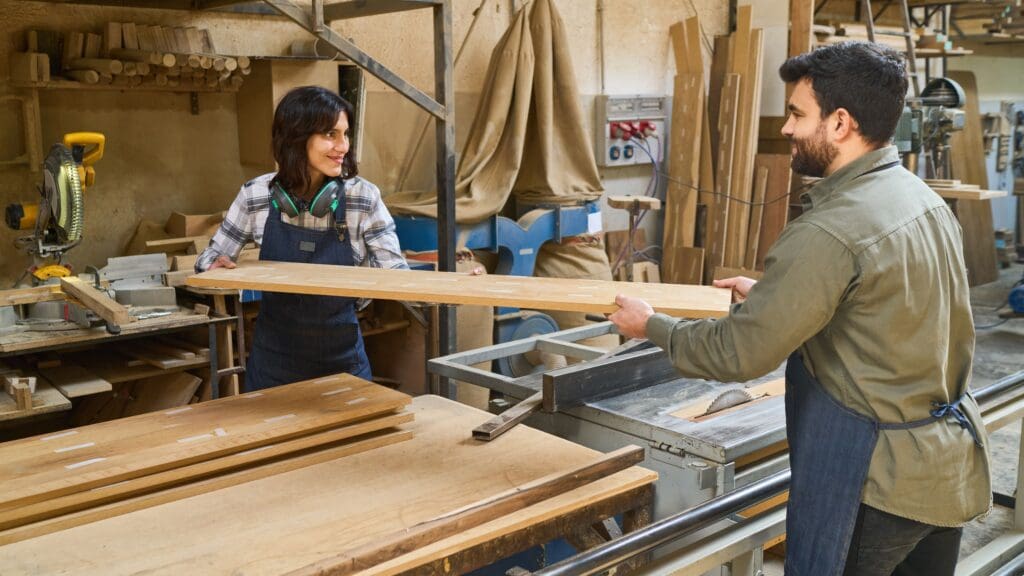 Two workers, a younger man and an older woman, collaborate in a bustling lumberyard