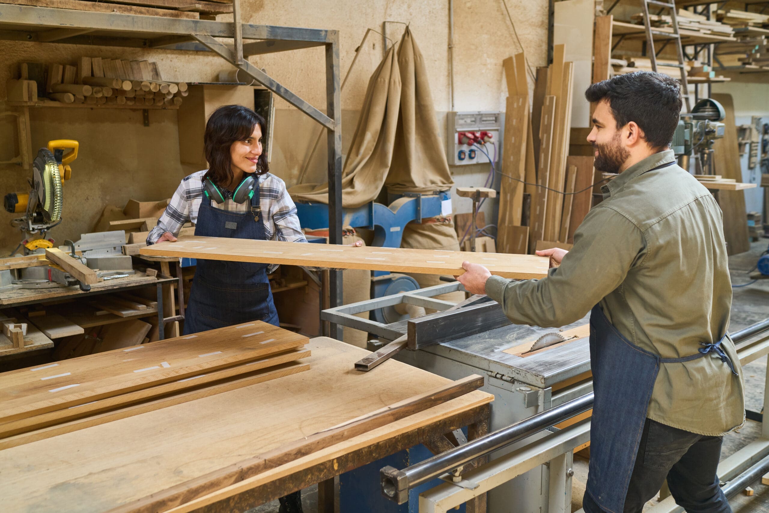 Two people working in a woodshop, holding a board between them and smiling.